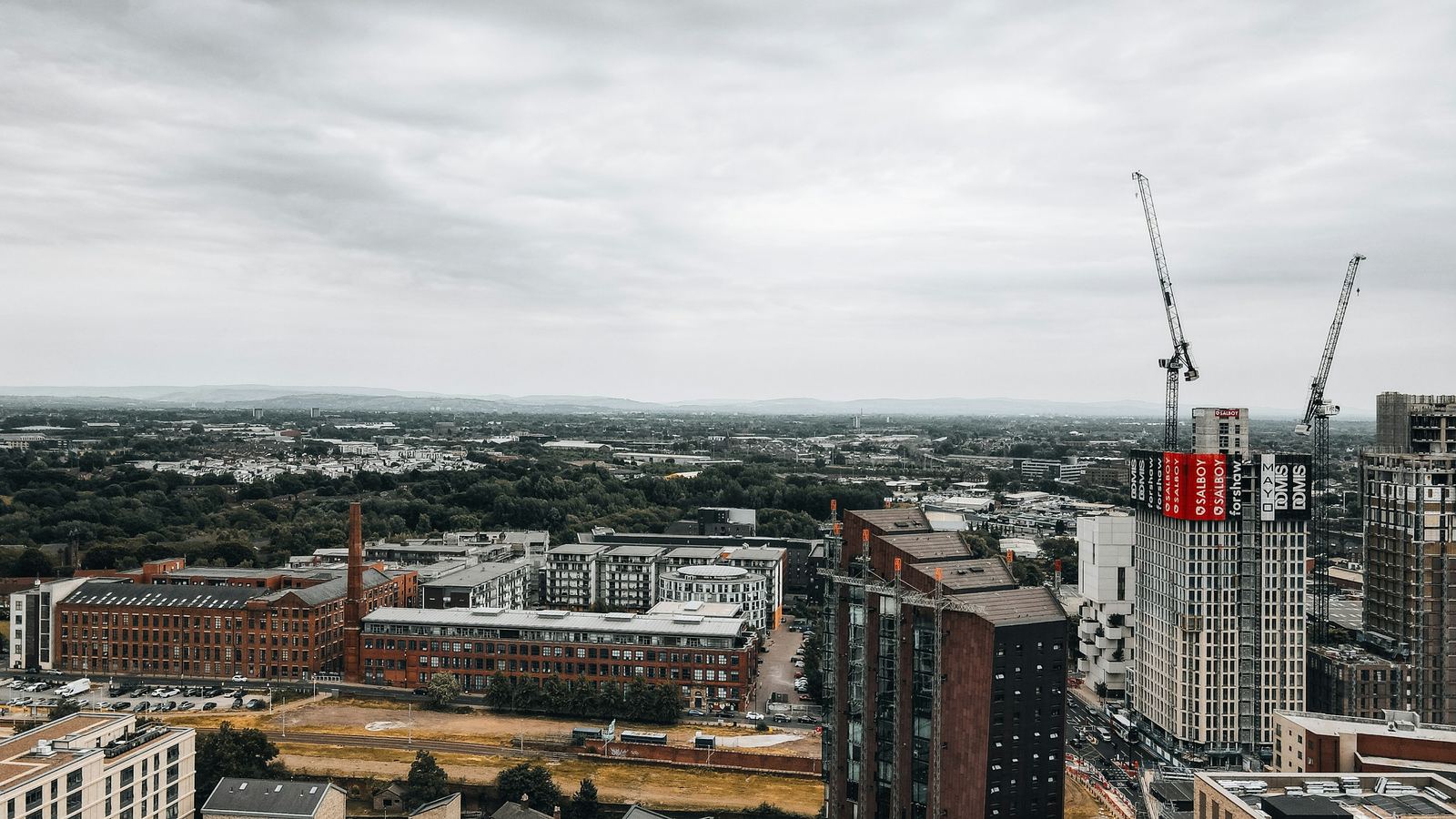 Tower cranes on new apartment blocks beside older brick mill buildings in Manchester, under an overcast sky