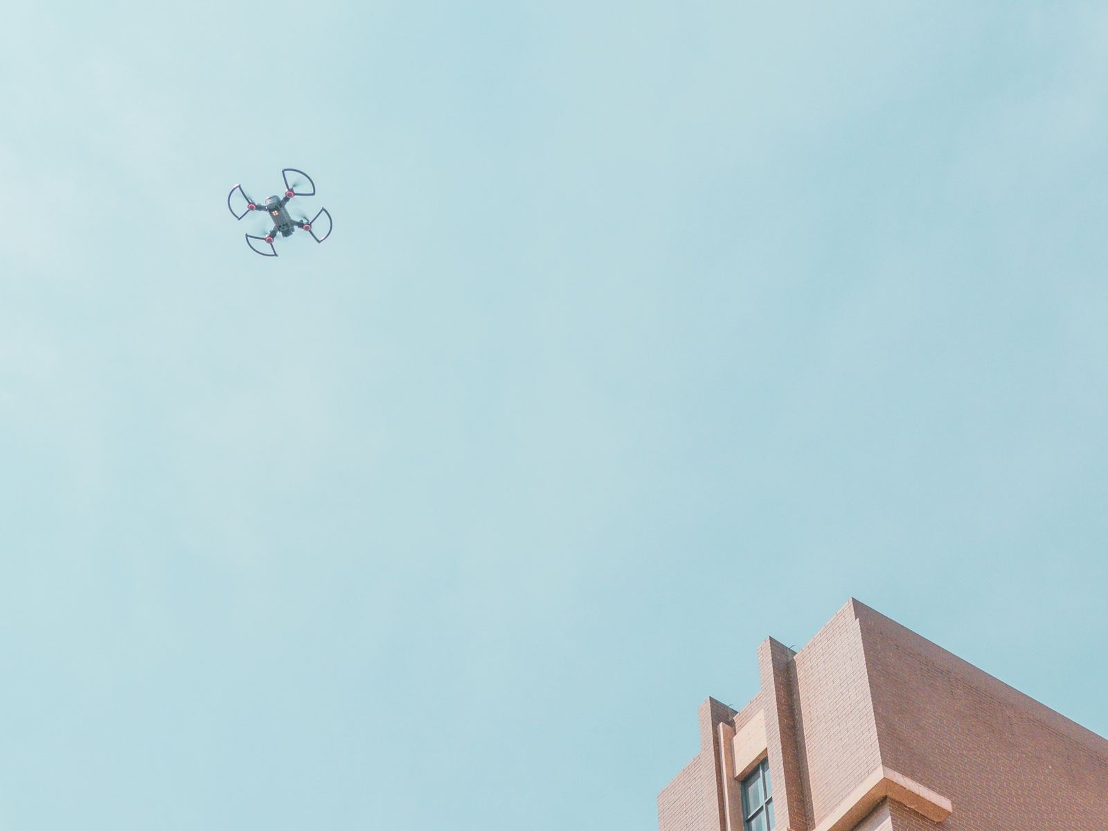A small drone flying in a clear sky above the brick parapet of a building