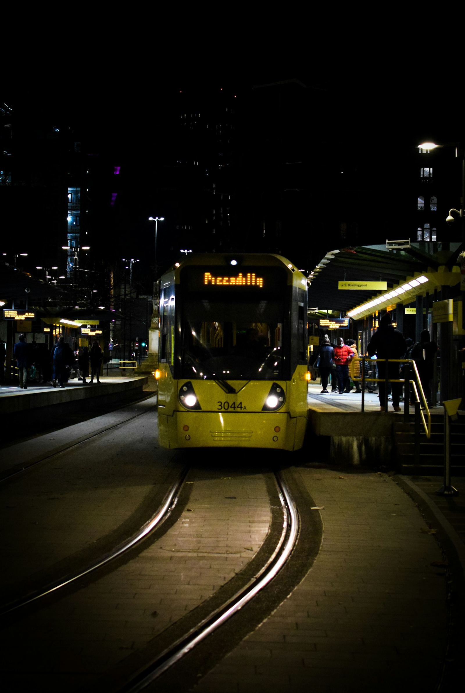 A yellow Metrolink tram bound for Piccadilly at a city centre stop at night