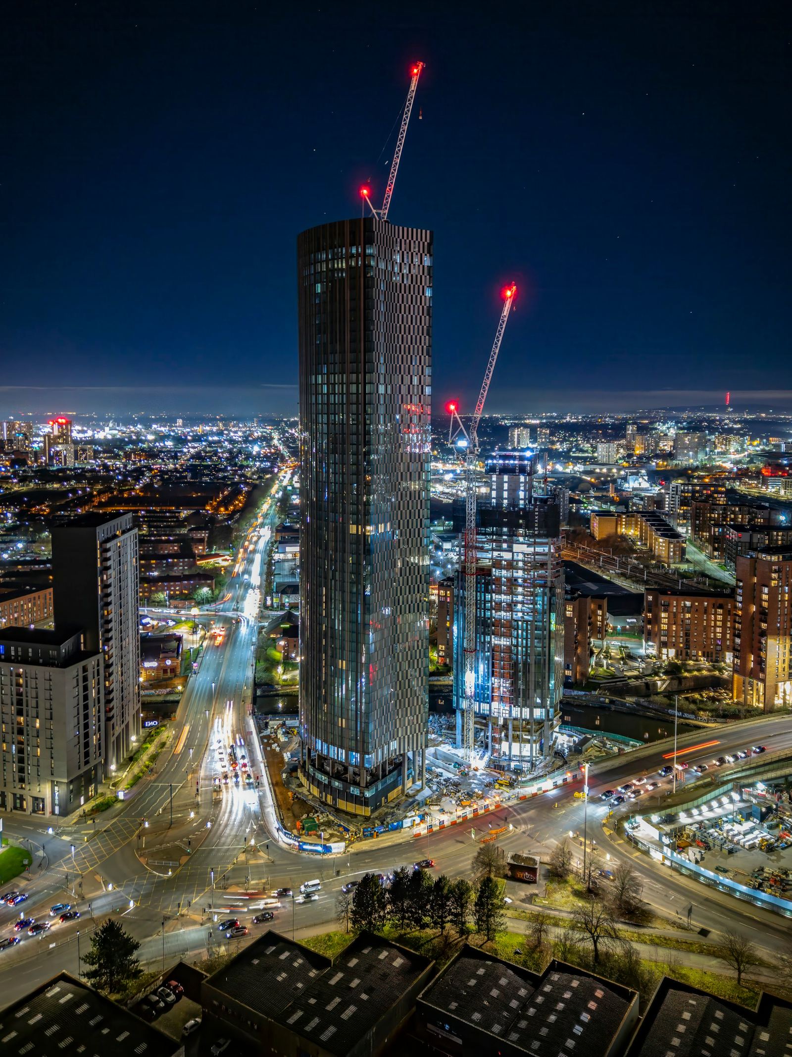 A residential tower under construction at night beside a busy junction, with two tower cranes lit by red aviation lights